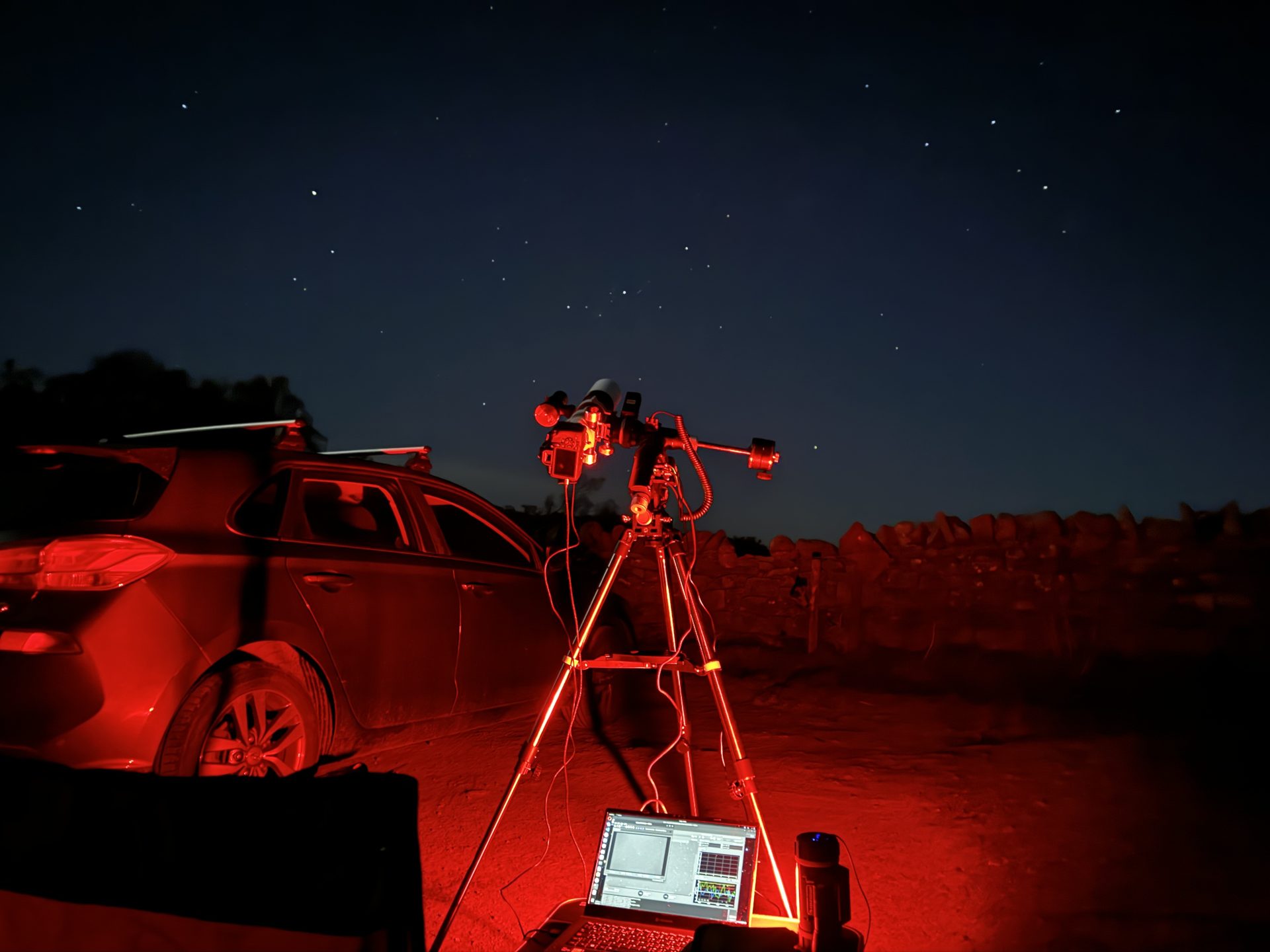 A telescope and camera set up under a starry sky, illuminated by red light